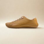 A imagem mostra um par de tênis casuais em couro feminino na cor bege, com acabamento texturizado e cadarço cinza em elástico. Os sapatos estão posicionados sobre uma superfície clara, valorizando seus detalhes minimalistas e design clean. O perfil é de corte baixo, com traseira acolchoada que sugere conforto.