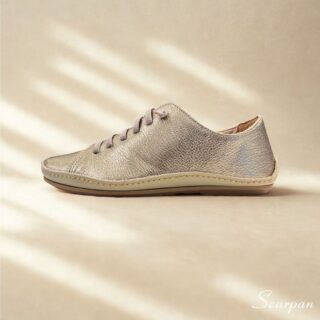 A imagem mostra um par de tênis casuais em couro feminino na cor prata envelhecido, com acabamento texturizado e cadarço cinza em elástico. Os sapatos estão posicionados sobre uma superfície clara, valorizando seus detalhes minimalistas e design clean. O perfil é de corte baixo, com traseira acolchoada que sugere conforto.