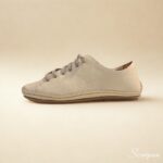 A imagem mostra um par de tênis casuais em couro feminino na cor off white, com acabamento texturizado e cadarço cinza em elástico. Os sapatos estão posicionados sobre uma superfície clara, valorizando seus detalhes minimalistas e design clean. O perfil é de corte baixo, com traseira acolchoada que sugere conforto.