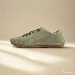 A imagem mostra um par de tênis casuais em couro feminino na cor verde, com acabamento texturizado e cadarço cinza em elástico. Os sapatos estão posicionados sobre uma superfície clara, valorizando seus detalhes minimalistas e design clean. O perfil é de corte baixo, com traseira acolchoada que sugere conforto.