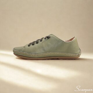 A imagem mostra um par de tênis casuais em couro feminino na cor verde, com acabamento texturizado e cadarço cinza em elástico. Os sapatos estão posicionados sobre uma superfície clara, valorizando seus detalhes minimalistas e design clean. O perfil é de corte baixo, com traseira acolchoada que sugere conforto.