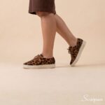 Tênis Feminino de Couro Pelo Onça Mia da Scarpan em ângulo de perfil em movimento. Modelo com estampa animal print de onça, cadarço marrom e solado Off White em TR, combinado com vestido marrom.