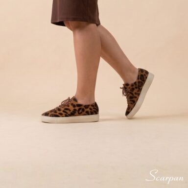 Tênis Feminino de Couro Pelo Onça Mia da Scarpan em ângulo de perfil em movimento. Modelo com estampa animal print de onça, cadarço marrom e solado Off White em TR, combinado com vestido marrom.