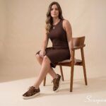 Modelo feminina sentada em cadeira de madeira usando o Tênis Feminino de Couro Pelo Onça Mia da Scarpan com vestido canelado marrom. Look casual elegante em tons terrosos.